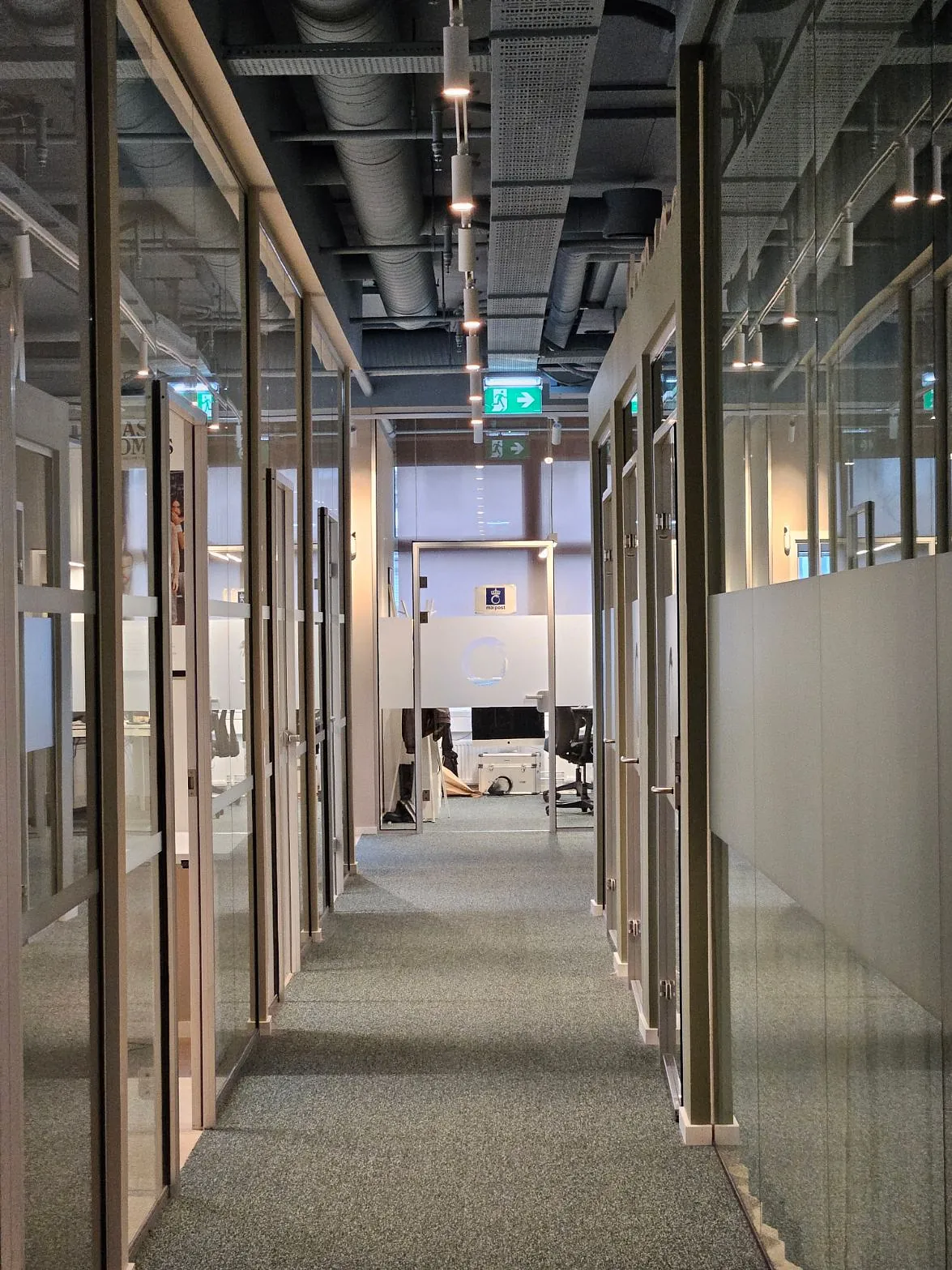 Glassveggede kontorer på begge sider av en moderne kontorgang med synlige ventilasjonsrør i taket.